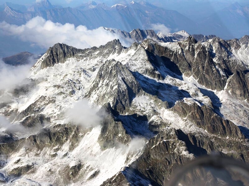 Panorama aérien de montagnes enneigées avec des sommets escarpés et des nuages flottants autour.