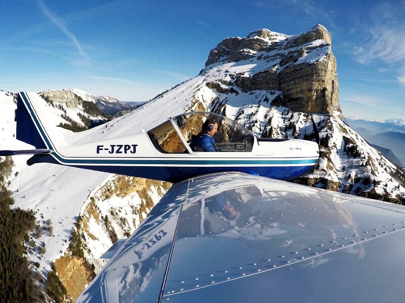 Petit avion léger en vol près d'une montagne rocheuse avec un pilote visible dans le cockpit.