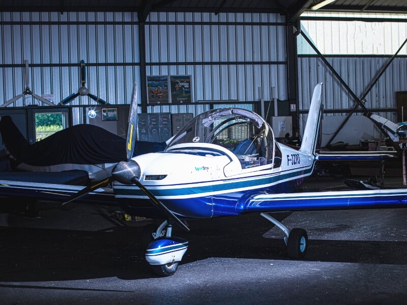 Avion léger bleu et blanc stationné dans un hangar d'aérodrome avec un intérieur sombre.