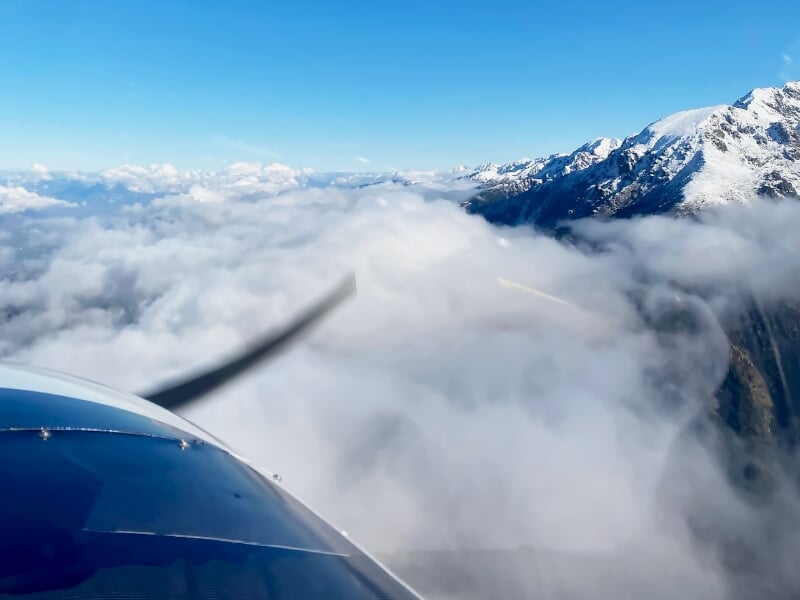 Vue depuis un avion en vol au-dessus des nuages avec des montagnes visibles à l'horizon.
