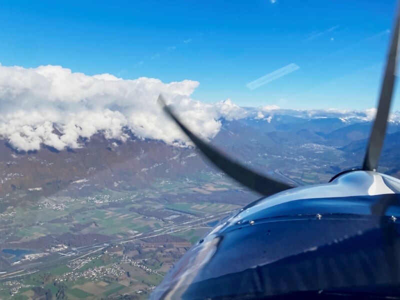 Vue en vol d'un avion léger au-dessus d'un paysage vallonné avec des nuages blancs.
