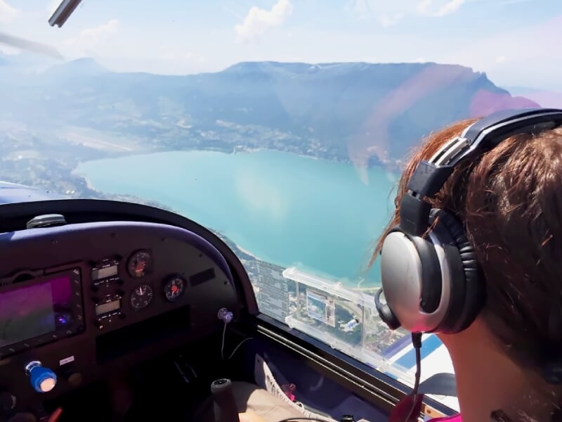 Pilote dans un petit avion léger survolant un lac turquoise entouré de montagnes verdoyantes.