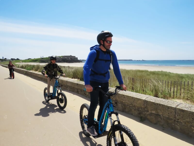 Groupe de cyclistes et de conducteurs de trottinettes électriques tout-terrain en balade sur un chemin en bord de plage, longeant une mer calme avec un fort en arrière-plan, sous un ciel bleu éclatant