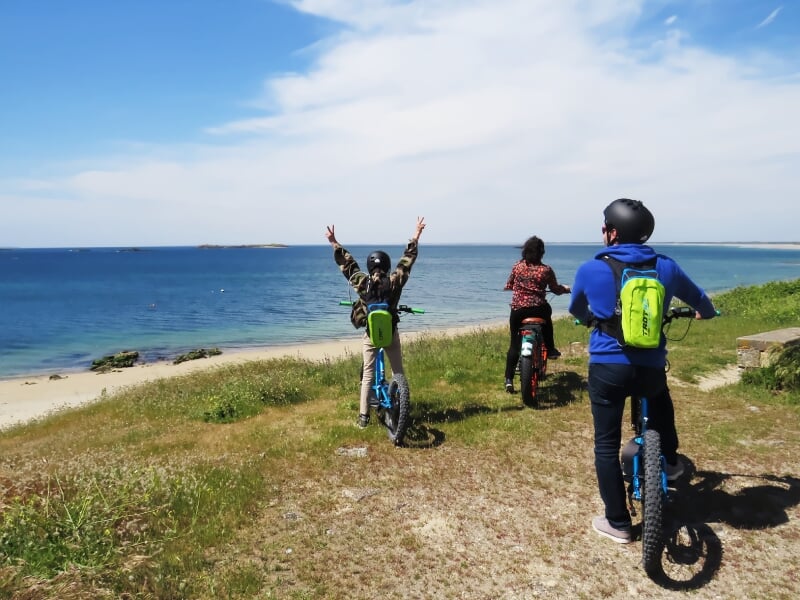 Trois personnes en trottinette électrique tout-terrain, équipées de casques et sacs à dos, admirant l’océan depuis un sentier côtier, avec une vue dégagée sur les îles au loin.