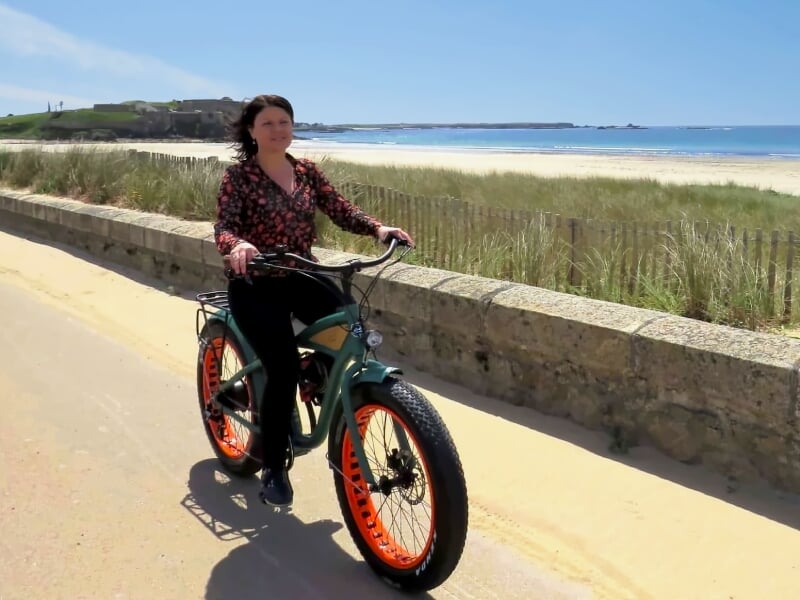 Femme en tenue décontractée roulant sur un vélo électrique vert avec pneus larges sur une route côtière ensoleillée.