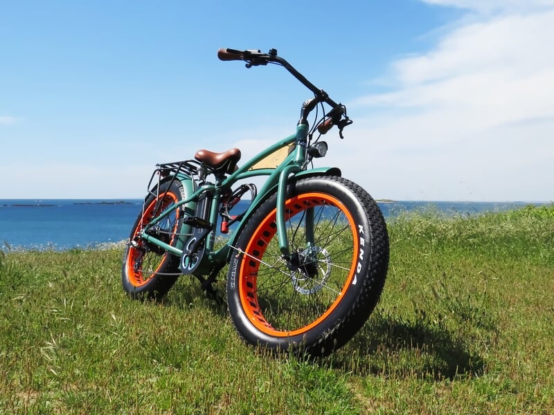 Vélo électrique vert avec pneus orange posé sur l'herbe face à la mer sous un ciel partiellement nuageux.