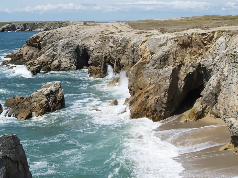 Falaises rocheuses surplombant une mer agitée avec des vagues s'écrasant sur la côte sous un ciel bleu clair.