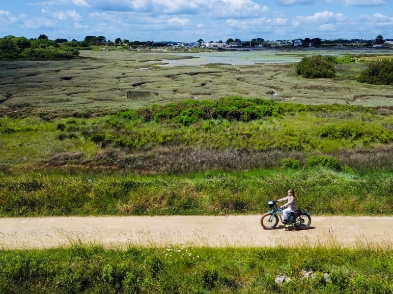 Femme à vélo sur chemin de terre bordé d’herbes vertes, marais verdoyant à l’arrière-plan, ciel bleu partiellement nuageux, maisons blanches visibles au loin.