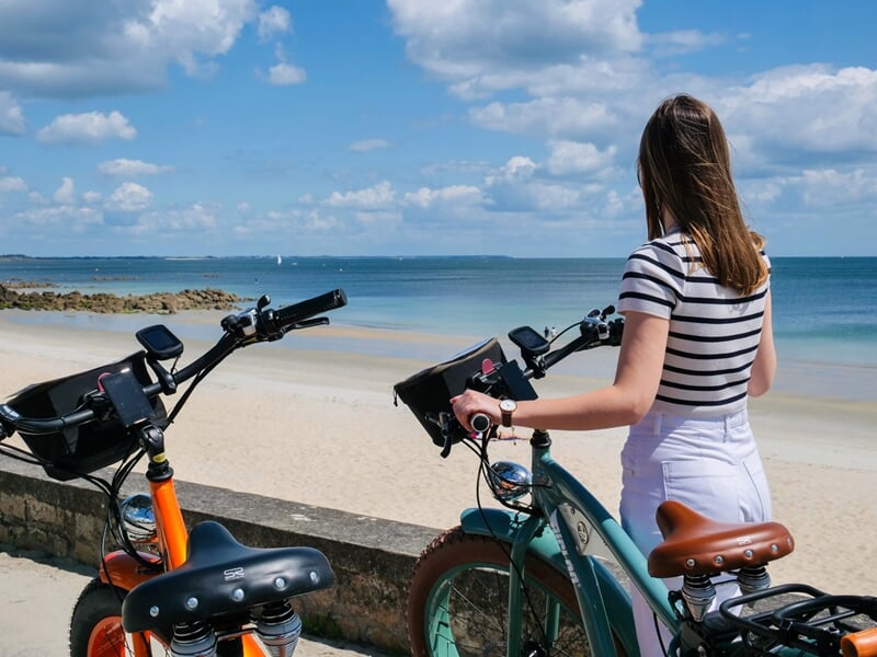 Femme en marinière et pantalon blanc tenant le guidon de sa trottinette électrique tout-terrain face à une plage de sable fin et une mer turquoise, avec un ciel partiellement nuageux
