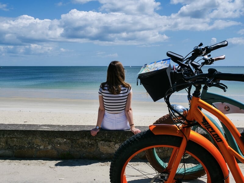 Femme assise sur un muret en bord de mer, regardant l'océan sous un ciel bleu parsemé de nuages. Devant elle, des trottinettes électriques tout-terrain aux couleurs vives stationnées sur le sable