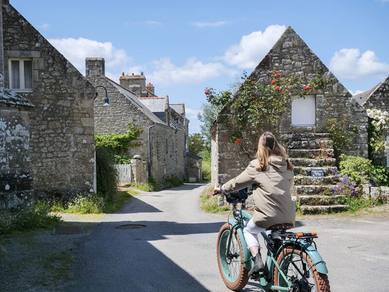 Personne à vélo électrique dans un village breton avec maisons en pierre sous un ciel bleu clair et quelques nuages.