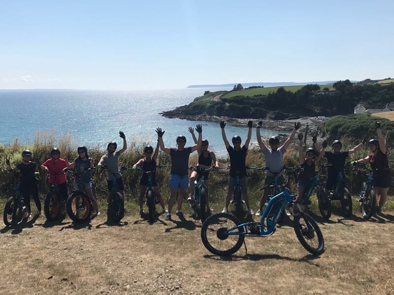 Groupe de personnes avec trottinettes électriques bleues posant au bord de la mer sous un ciel clair et ensoleillé.