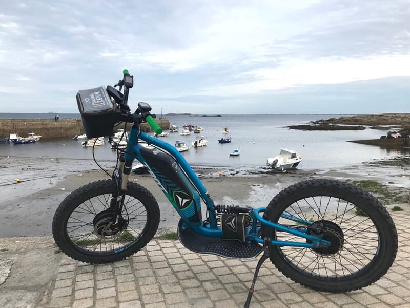 Trottinette électrique bleue garée sur un quai pavé avec des bateaux et la mer en arrière-plan sous un ciel nuageux.