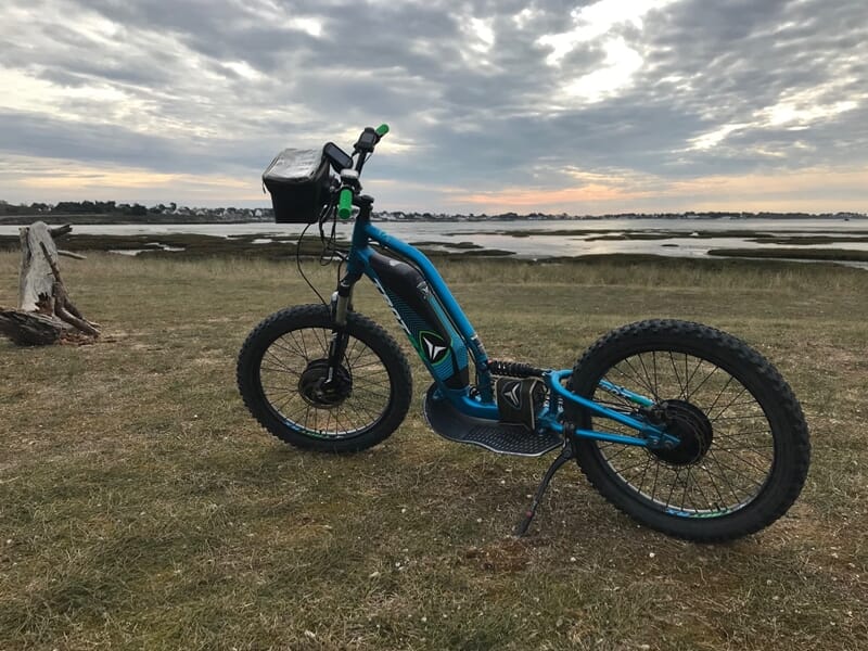Trottinette électrique bleue posée sur une pelouse avec vue sur la mer et un ciel nuageux en fin de journée.