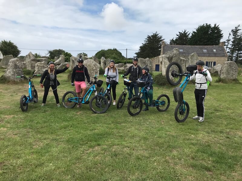 Groupe de personnes avec trottinettes électriques bleues posant devant un site de pierres dressées sur une pelouse verte.