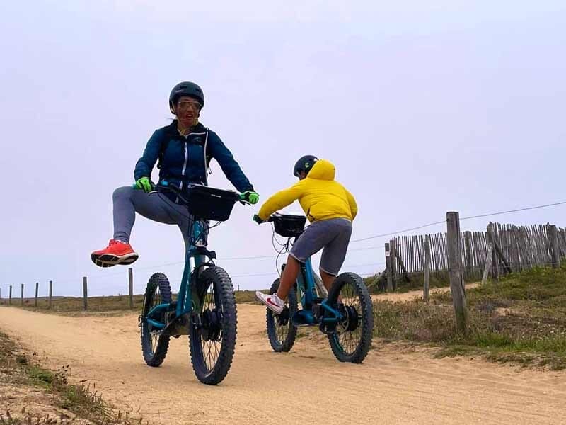 Deux personnes en tenue sportive avec trottinettes électriques bleues roulant sur un chemin de terre en bord de mer.