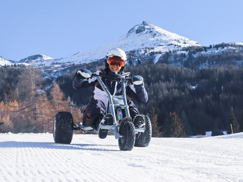 Personne souriante sur un kart à trois roues sur une piste enneigée avec montagnes en arrière-plan.