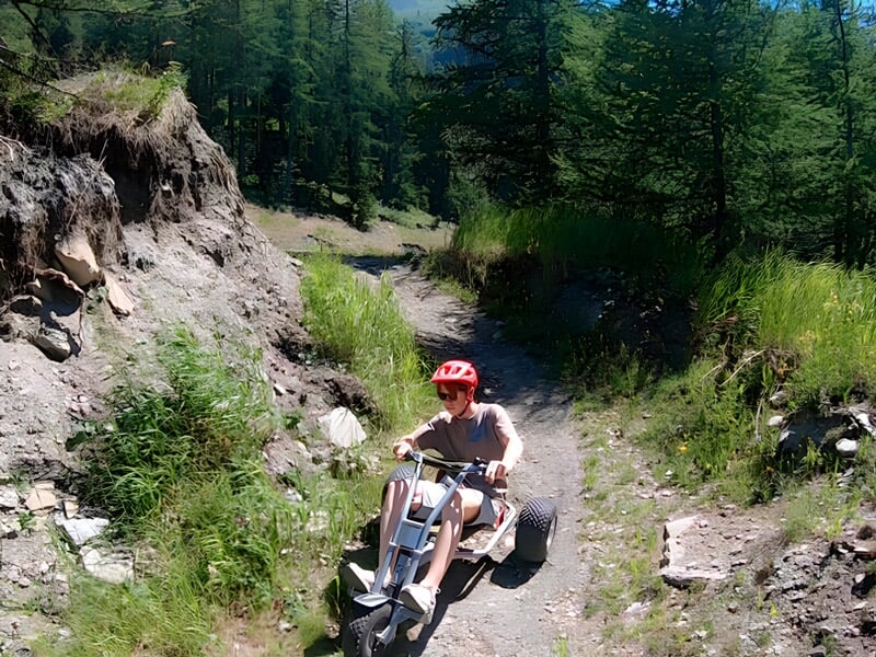 Enfant portant un casque rouge conduisant un kart tout-terrain sur un chemin de terre en forêt.