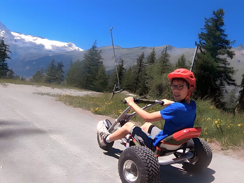 Jeune garçon souriant portant un casque rouge conduisant un kart tout-terrain sur une route de montagne.