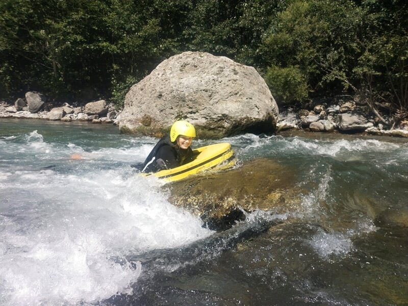 Personne en combinaison noire et casque jaune glissant en hydrospeed sur une rivière avec des rochers.