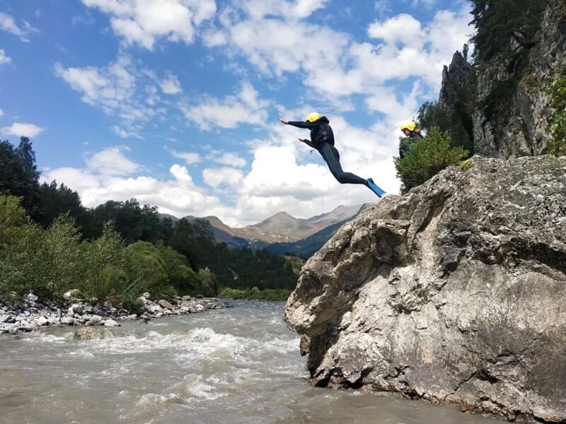 Individu sautant d'un rocher dans une rivière entourée de forêt et montagnes en arrière-plan.