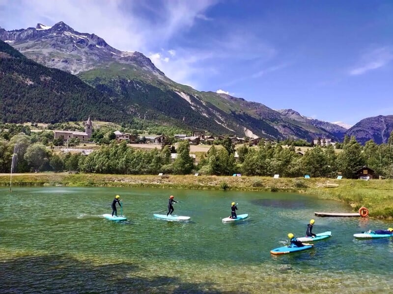 Personnes debout sur des planches de stand-up paddle sur un lac calme avec montagnes en arrière-plan.