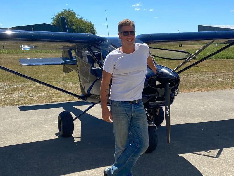 Un homme souriant en t-shirt blanc et jean pose devant un petit avion léger sur une piste ensoleillée.