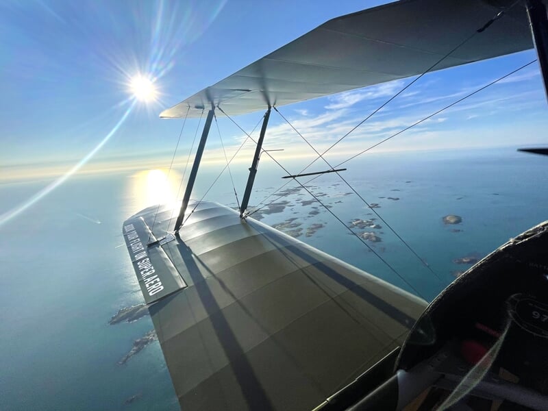 Vue depuis le poste de pilotage d'un biplan en vol, montrant une des ailes et un paysage côtier avec des îlots dispersés sur une mer calme, avec le soleil brillant dans un ciel bleu clair