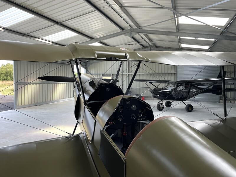 Intérieur d'un hangar à avions montrant un biplan classique au premier plan avec d'autres avions en arrière-plan, éclairé par une lumière naturelle