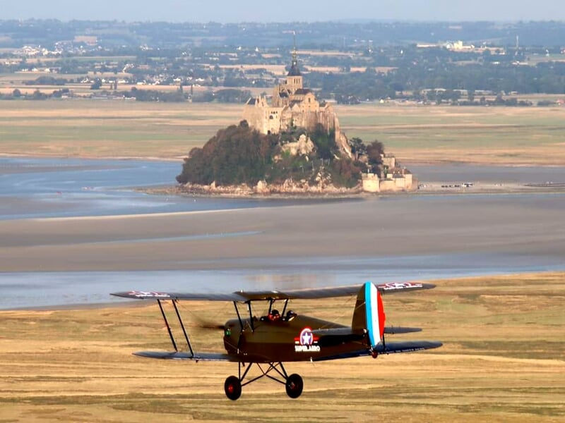 Un biplan classique en vol devant le Mont-Saint-Michel, avec son reflet dans l'eau à marée basse et une vaste étendue de plages et de champs en arrière-plan