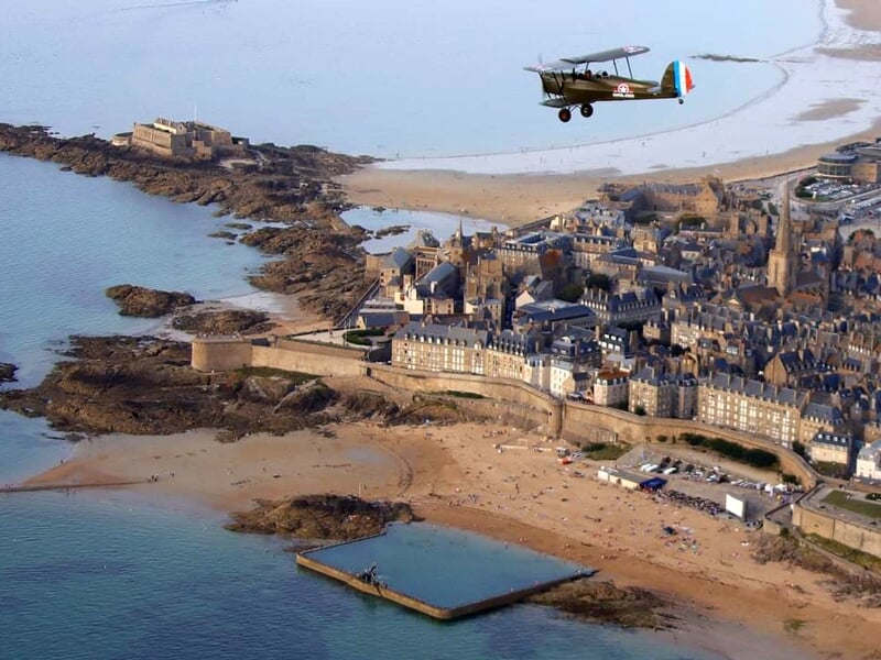 Vue aérienne d'un biplan classique survolant Saint-Malo avec ses plages et ses fortifications, ses bâtiments historiques et la mer formant un paysage pittoresque