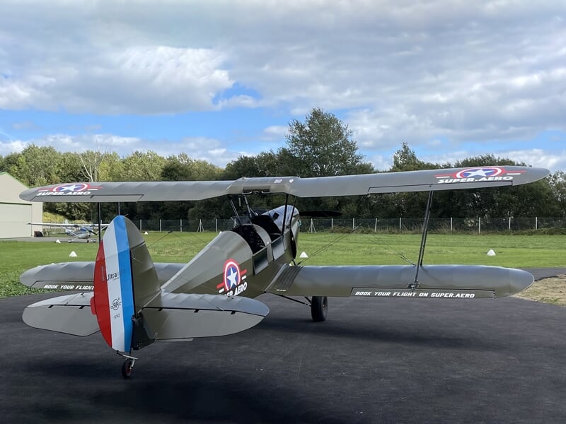 Biplan classique au sol devant un hangar, avec des ailes aux couleurs nationales et une hélice en bois, sur un tarmac entouré d'herbe