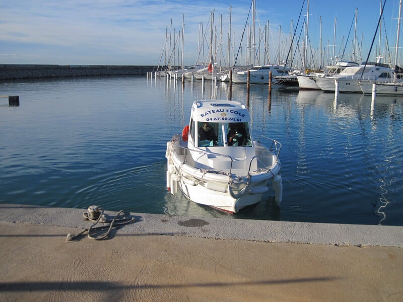 Petit bateau blanc vu de face amarré au quai, avec plusieurs voiliers alignés en arrière-plan dans le port.