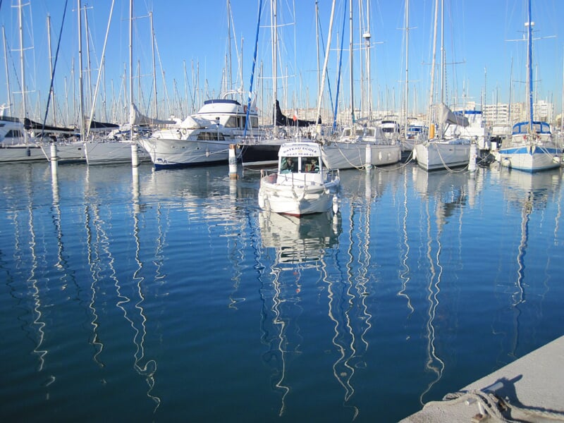 Bateau blanc au centre d'un port avec de nombreux voiliers amarrés, reflet des mats dans l'eau calme sous un ciel clair.