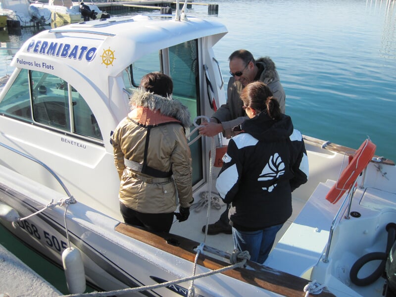 Trois personnes discutent sur un bateau blanc amarré au port, sous un ciel bleu avec des voiliers en arrière-plan.
