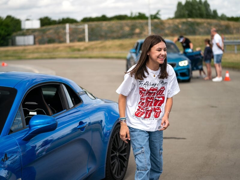 Jeune fille souriante, vêtue d'un t-shirt des Rolling Stones et d'un jean décontracté, marchant à côté d'une voiture de sport Alpine bleue garée sur une piste. En arrière-plan, plusieurs personnes son