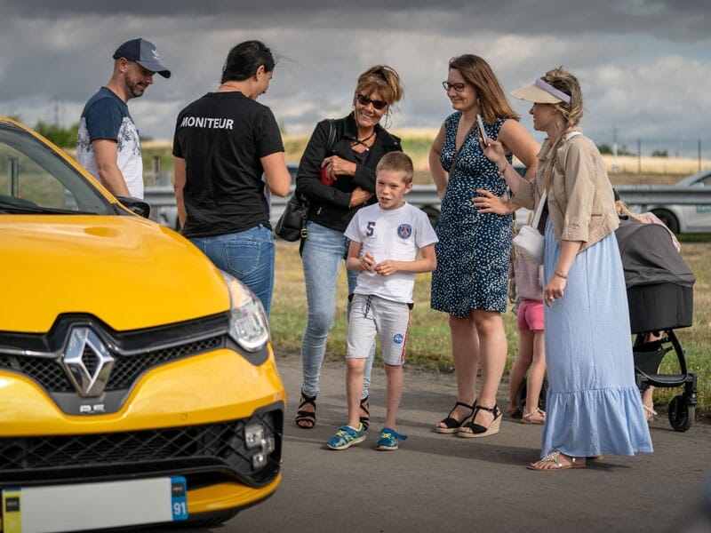 Petit groupe de personnes rassemblées autour d'une Renault Clio jaune. Une monitrice portant un t-shirt noir semble donner des instructions. Un jeune garçon vêtu d'un t-shirt du PSG est au centre de l