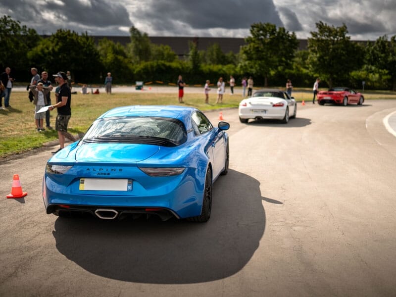 Voiture de sport Alpine A110 bleue sur une piste, suivie par deux autres voitures. La scène se déroule sous un ciel partiellement couvert, avec quelques spectateurs au loin près de cônes de signalisat