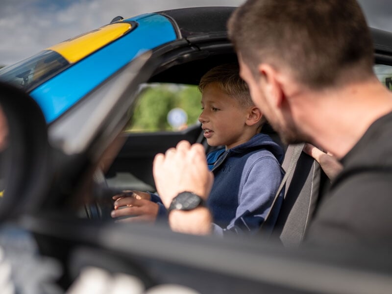 Un jeune garçon, assis dans une voiture de sport, est attentif aux conseils donnés par un homme assis à côté de lui. La scène se déroule dans l'habitacle, où l'homme semble lui expliquer les commandes