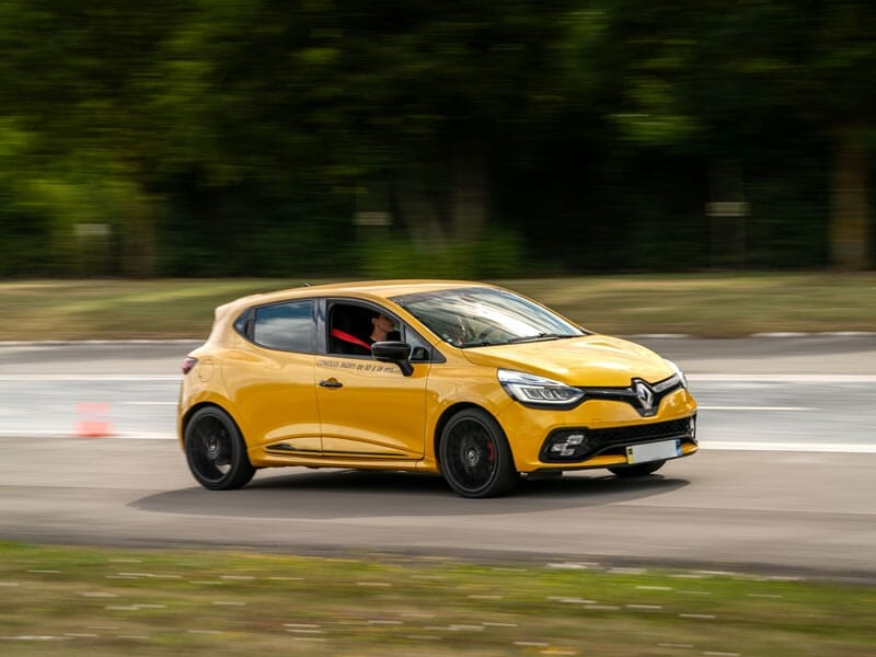 Renault Clio jaune vif en pleine vitesse sur une piste, capturée avec un effet de flou montrant le mouvement. Le conducteur, concentré, porte une tenue rouge visible à travers la fenêtre ouverte. La v