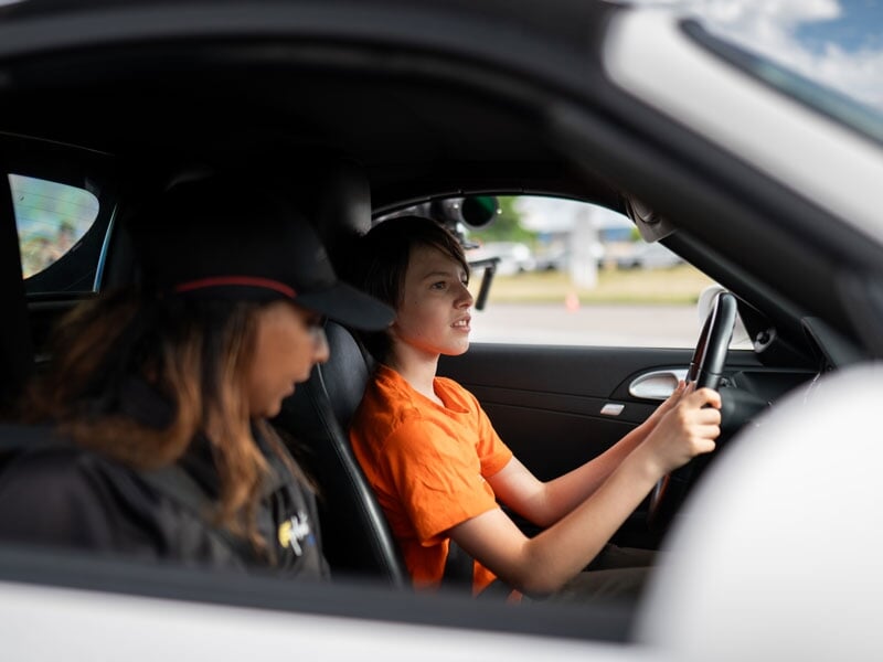 Jeune garçon au volant d'une voiture de sport, concentré, vêtu d'un t-shirt orange. À côté de lui, une femme portant une casquette noire semble observer ou donner des instructions. La scène se déroule