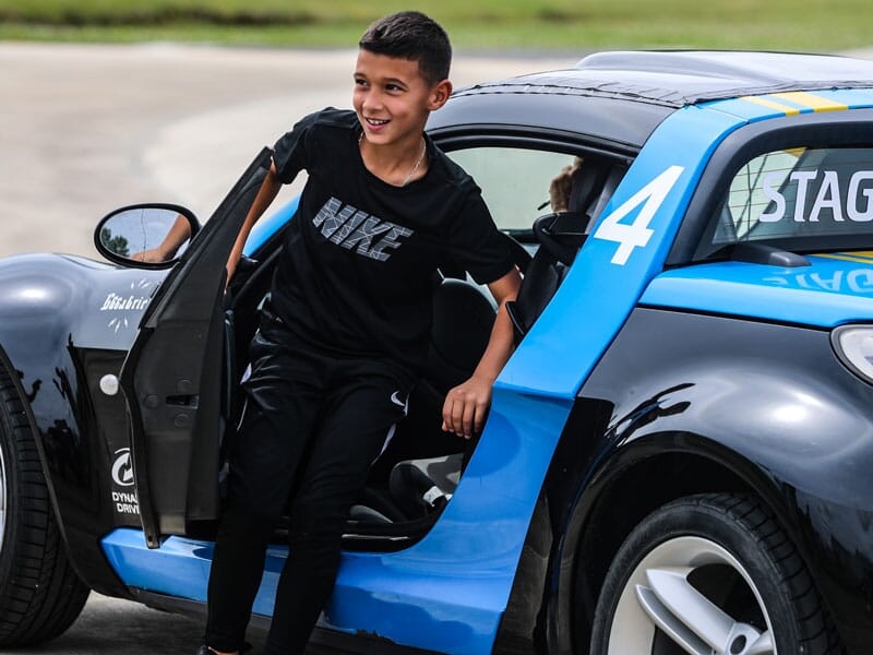 Garçon sortant d'une voiture de sport noire et bleue, le visage illuminé par un grand sourire. Il porte un ensemble sportif noir et la porte de la voiture est ouverte, capturant l'excitation après une