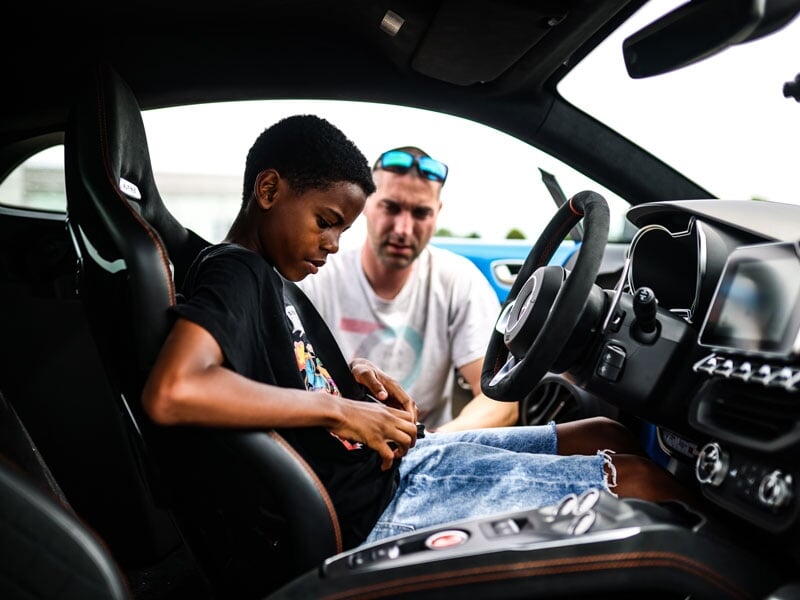 Adolescent assis dans une voiture de sport, ajustant sa ceinture de sécurité, tandis qu'un homme en arrière-plan lui donne des indications. La scène montre un moment d'apprentissage avant une session 