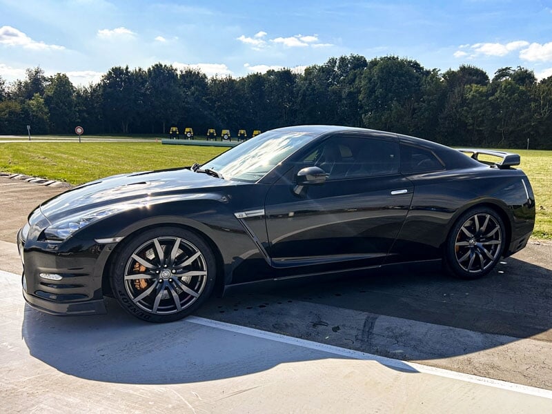 Nissan GT-R noire, vue avant, carrosserie brillante sous le soleil. Ligne sportive et puissante, entourée de pelouse et d’arbres, dans un cadre naturel sur circuit