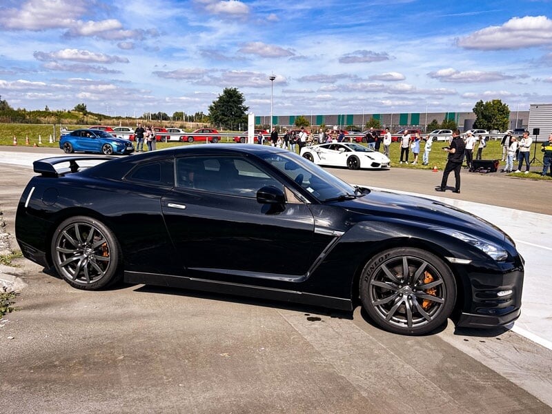 Nissan GT-R noire, profil bas et jantes foncées, garée sur circuit. En arrière-plan, foule et autres voitures sportives, ambiance de rassemblement automobile sous un ciel dégagé