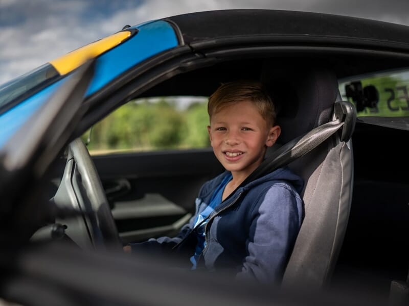 Jeune garçon souriant assis au volant d'une voiture de course, prêt à piloter sur un circuit automobile.