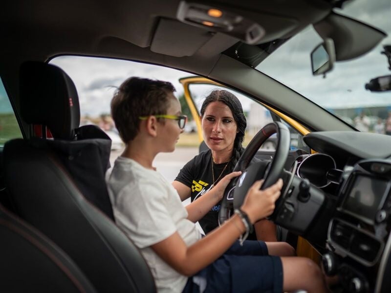 Une femme explique à un garçon assis au volant d'une voiture les bases du pilotage sur un circuit automobile.