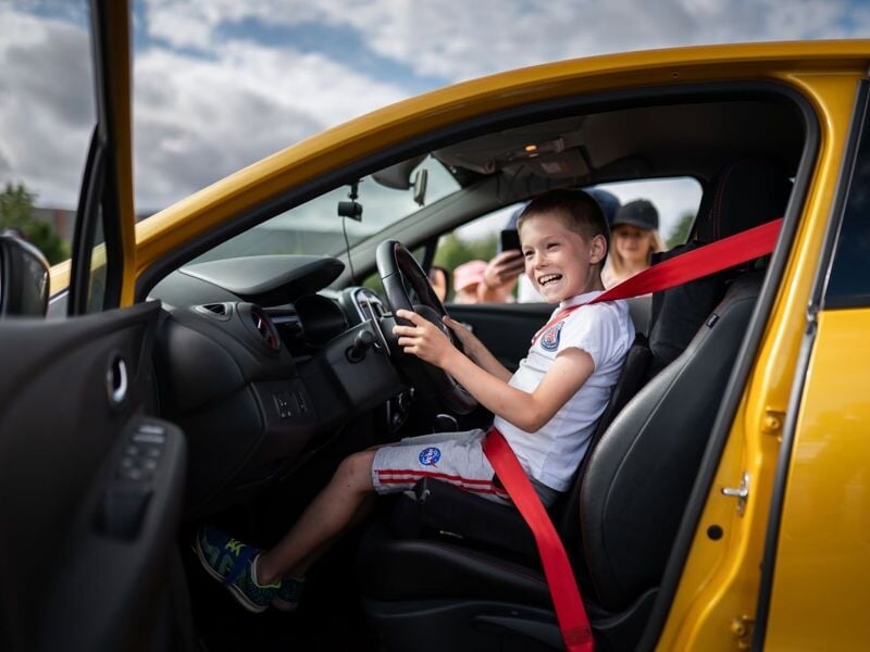 Un jeune garçon souriant est assis au volant d'une voiture jaune, attaché avec une ceinture de sécurité rouge.
