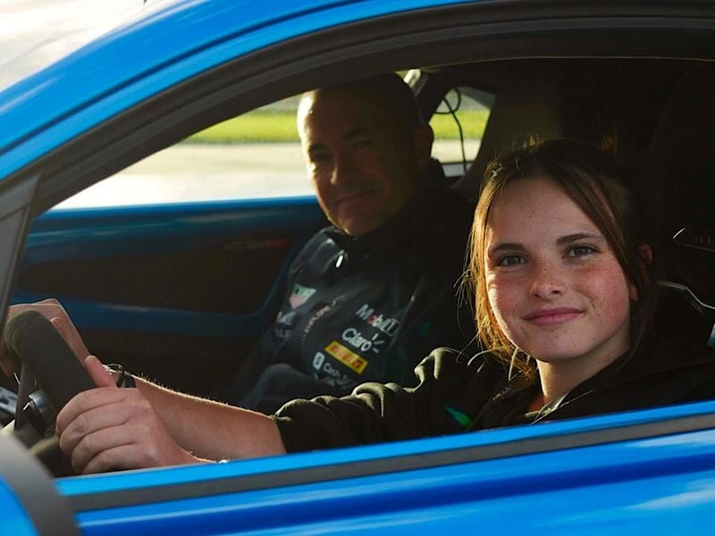 Une jeune fille souriante est assise au volant d'une voiture bleue avec un homme à côté, tous deux prêts à conduire.