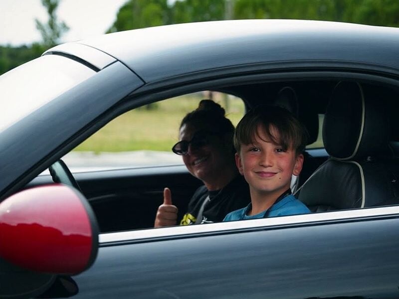 Un garçon souriant est assis dans une voiture grise avec une femme à côté qui fait un pouce levé en signe d'encouragement.